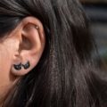 Close-up of a person’s ear wearing two black enamel cat stud earrings made of sterling silver, with dark hair framing the ear.