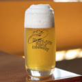 A Burger Eyes Tumbler filled with light, frothy beer sits on a wooden table. The glass features the "Highland Park Brewery" logo with a cowboy hat illustration, while the softly blurred background glows in warm tones.