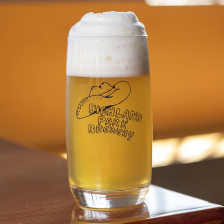 A Burger Eyes Tumbler filled with light, frothy beer sits on a wooden table. The glass features the "Highland Park Brewery" logo with a cowboy hat illustration, while the softly blurred background glows in warm tones.