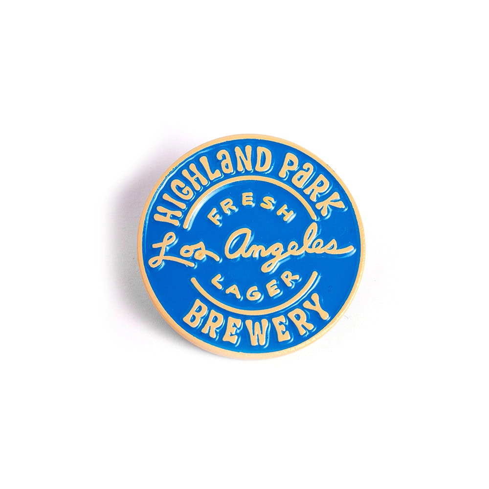 A round blue pin with yellow text that reads, “Highland Park Brewery Fresh Los Angeles Lager,” in a mix of print and cursive lettering.