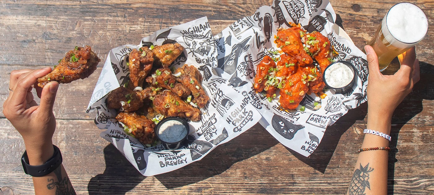 Two hands reach in from either side of a rustic wooden table at Highland Park Brewery Los Angeles, one holding a chicken wing, the other a glass of beer. On the table are baskets of saucy wings and cups of ranch—classic food and beer menu pairings.