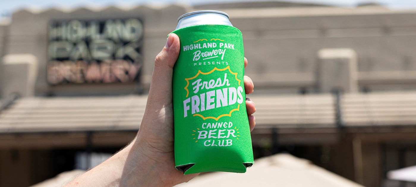 A hand holds a can in a bright green koozie that reads "Highland Park Brewery Presents Fresh Friends Canned Beer Club," highlighting their exclusive beer membership, with a blurred building and umbrellas in the background.