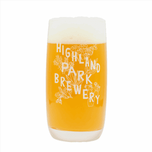 A glass of hazy golden beer with a foamy head is poured into the Native Plants Glass, featuring white floral illustrations and "Highland Park Brewery" printed on it, set against a plain white background.