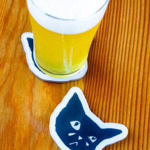 A glass of light beer rests on a Sad Cat Coaster (Set of 4) featuring a black cat face, with another matching coaster beside it, both placed on a wooden surface.