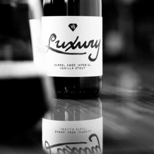 A close-up of a bottle labeled "Luxury Barrel Aged Imperial Vanilla Stout" with its reflection on a glossy surface, captured in black and white—a tempting scene for any beer membership enthusiast. Another blurred bottle appears on the left side.