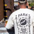 A person outdoors holds a drink while wearing the Owl Long Sleeve—a white shirt with bold black graphics, “Highland Park Brewery” text, and moon and owl illustrations on the back.