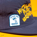 A Sad Cat Baseball League Pin (2026) rests on a bright yellow surface, featuring a patch of a baseball player pitching and "Highland Park Brewery Baseball Club" label, with "Highland Park Brewery" in yellow embroidery on navy.