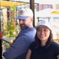 A man and woman stand back to back, smiling outdoors in bright patio setting with umbrellas. Both wear light-colored HPB LA baseball caps; the man in a blue shirt holds a drink, while the woman wears a black shirt.