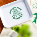 The HPB Turple Rolling Tray, featuring a white base with a green hop cone illustration and "Highland Park Brewery" text, rests on fabric beside blurred green items and a partially visible graphic.