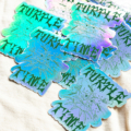 A 2-pack of Turple Time Stickers featuring bold stylized “PURPLE TIME” text. Holographic finish reflects green, blue, and purple shades. Displayed on a light fabric surface.