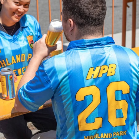 Two people sit at an outdoor table, both wearing blue HPB Soccer Jerseys. One, facing away, holds a glass of beer; the other smiles and holds a beverage can. An orange barrier appears in the background.