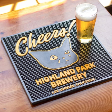 A glass of beer rests on the HPB Sad Cat Cheers! Bar Mat, which features a cartoon cat, "Cheers!", and "Highland Park Brewery, Los Angeles, California," all atop a wooden table.